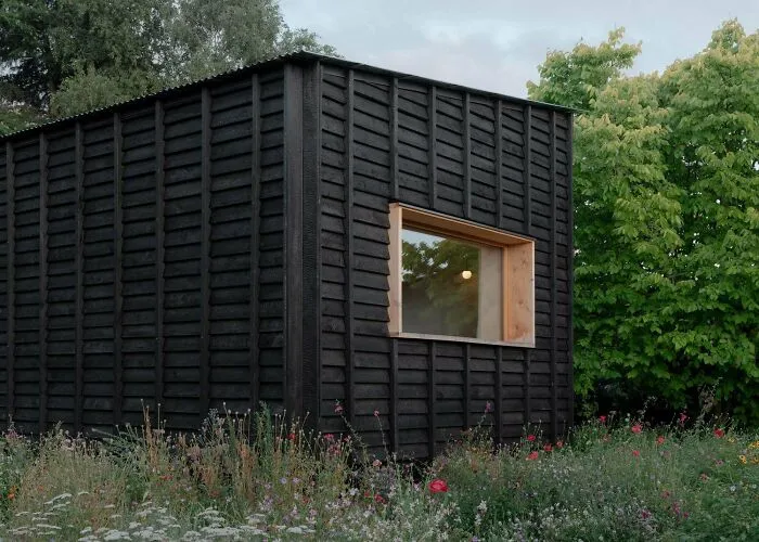 A minimalist black wooden cabin with a single large window surrounded by a lush wildflower garden.