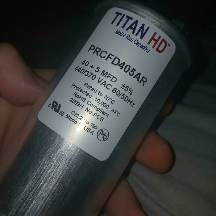 A hand holding a Titan HD motor run capacitor as part of frugal tips to fix household items.