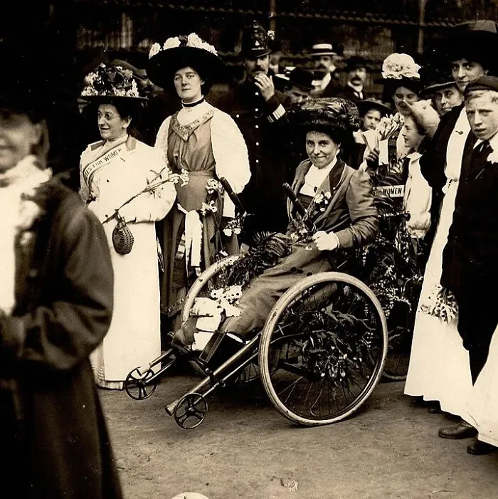 A historical gathering of women, including a woman in a decorated wheelchair, surrounded by other women and men.