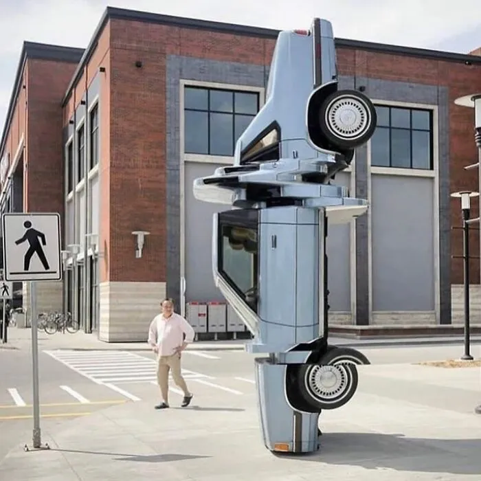A unique street art installation featuring a light blue car standing vertically on the pavement in an urban setting.