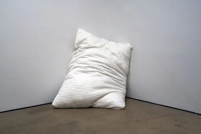 A large white pillow leaning against a corner of a gallery room as part of unusual sculptures