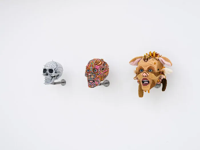 Three unusual sculptures displayed on a wall starting from a white skull to a detailed plush face