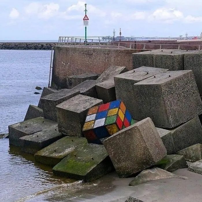 An artistic street art display of a giant Rubik's cube among concrete blocks near the ocean.