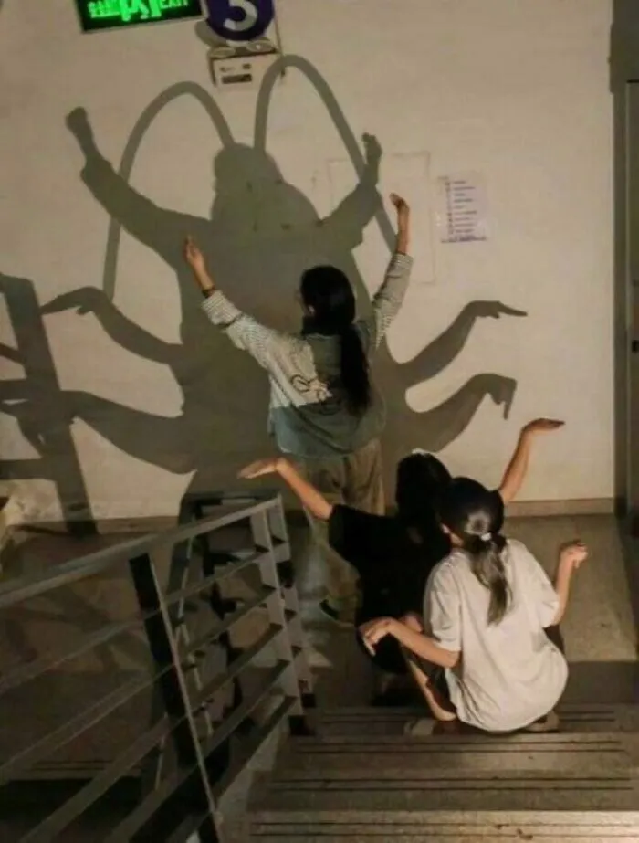 Unhinged image of three people creating a giant insect shadow on a wall, arms and legs forming antennae and limbs.