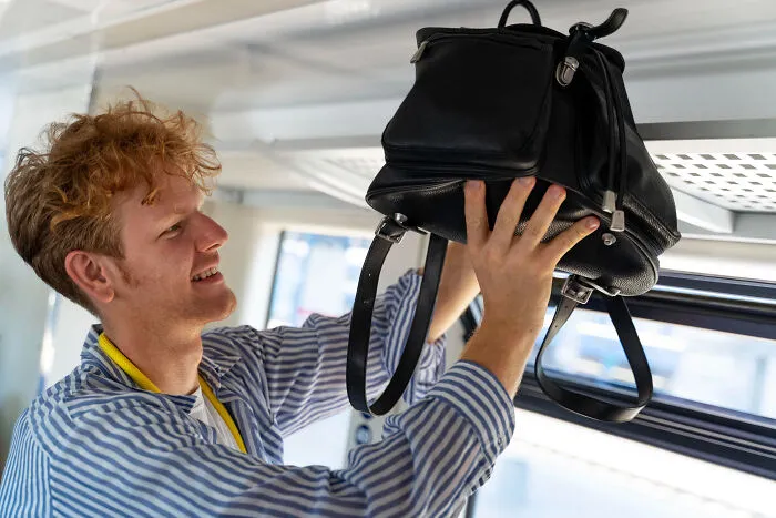 A young man placing his black backpack in the overhead bin on a flight next to his plane seatmate.
