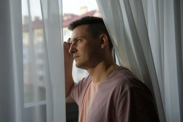 A thoughtful man looking out of a window behind sheer curtains, contemplating ridiculous arguments.