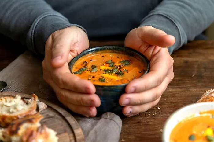 Person holding a bowl of pumpkin soup garnished with seeds and herbs