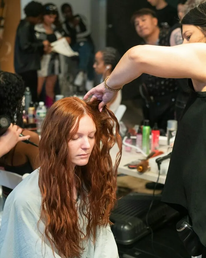A woman with long red hair getting her hair styled, with other people in the background, a glimpse into Hollywood life.