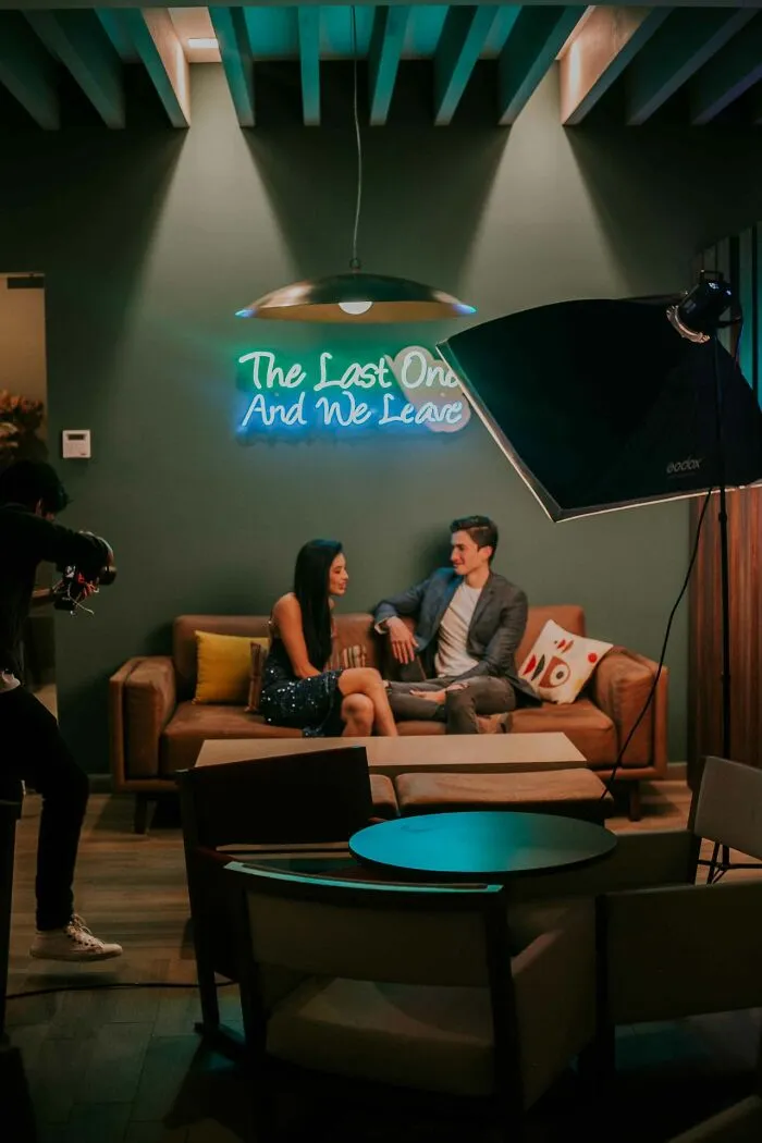 A couple sitting on a couch under a neon sign that reads The Last One And We Leave, hinting at Hollywood rumors.