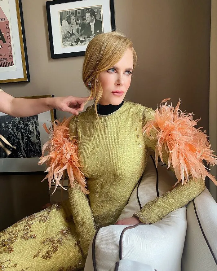 Nicole Kidman in a golden dress with feathered sleeves, looking off to the side, revealing Hollywood secrets.