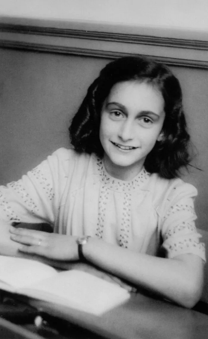 Anne Frank, an iconic young woman in history, smiling while sitting at a table with an open book.