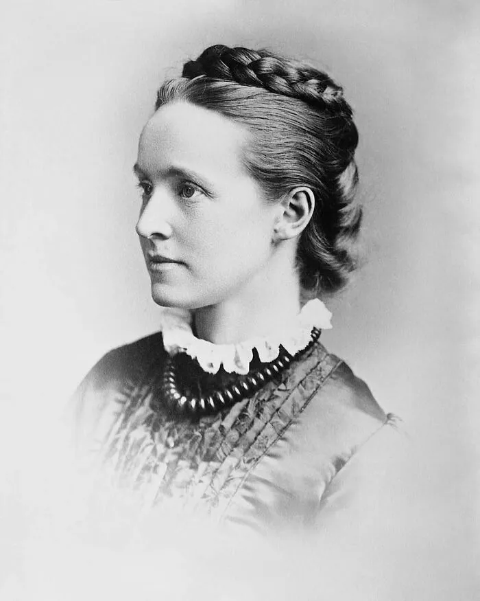 Profile black and white portrait of a young woman with braided hair and a ruffled necklace, a figure in women in history.