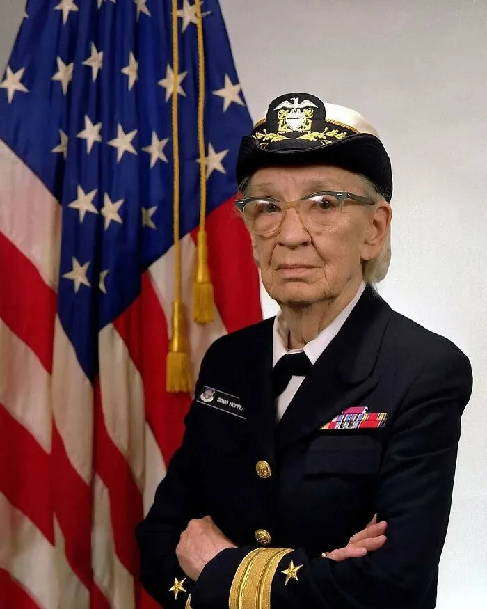 An older woman in a US Navy uniform with glasses, standing in front of an American flag, representing women in history.