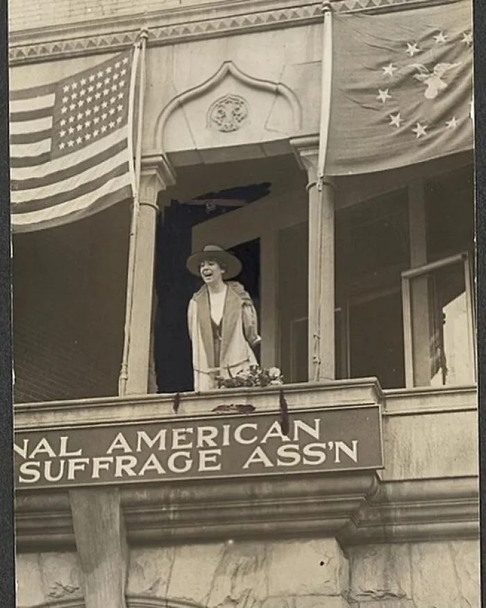 Woman on a balcony with US flags, celebrating a moment for Women In History, possibly suffrage.