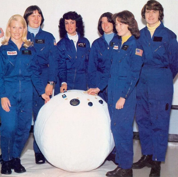 Six women in blue NASA jumpsuits stand around a large white spherical object, representing Women in History.