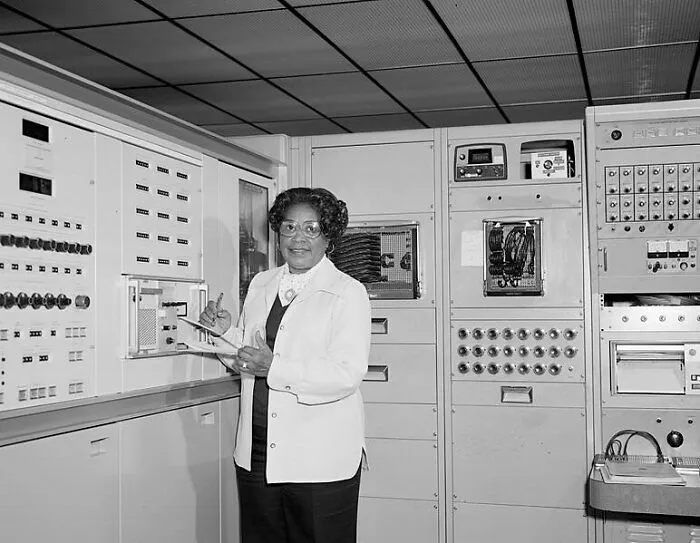 An African American woman in history stands confidently beside complex computer equipment, holding a clipboard.