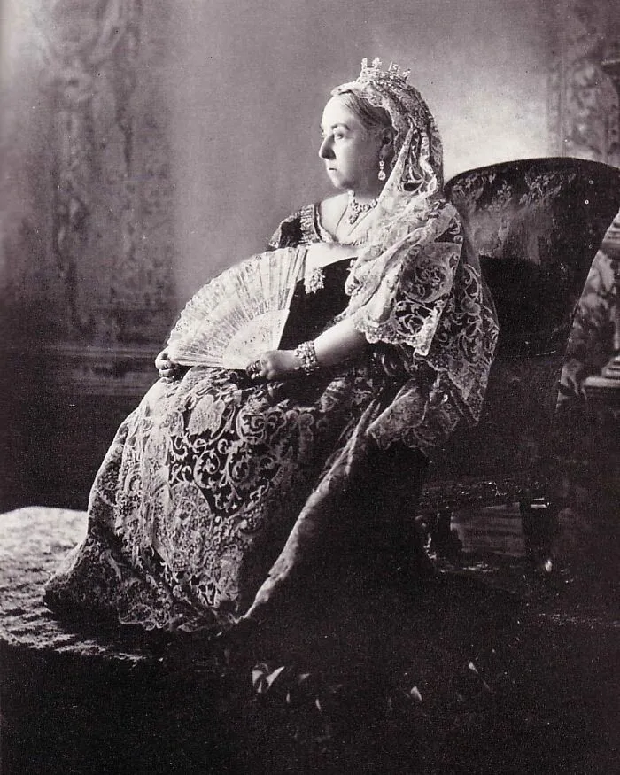 A regal black and white photo of Queen Victoria, an iconic figure among Women in History, seated with a fan, adorned in a formal gown and tiara.