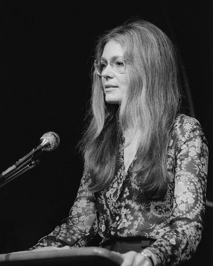 A woman with long hair and glasses sits at a keyboard with a microphone, a key figure in Women in History.