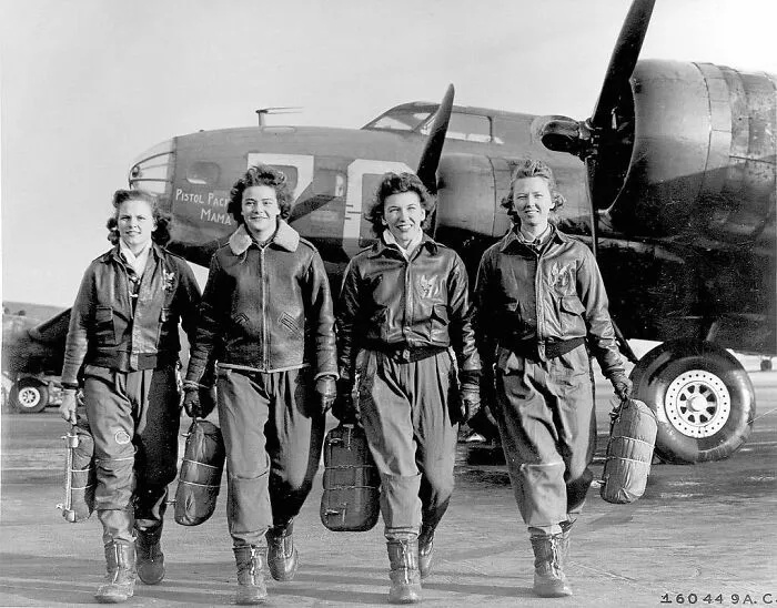Four female pilots, important Women in History, in flight suits and bomber jackets, walking across an airfield with a B-17 bomber in the background.