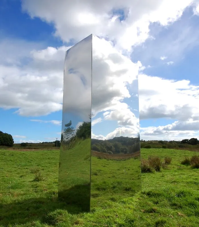 Tall reflective monolith sculpture standing on green grass under a partly cloudy sky