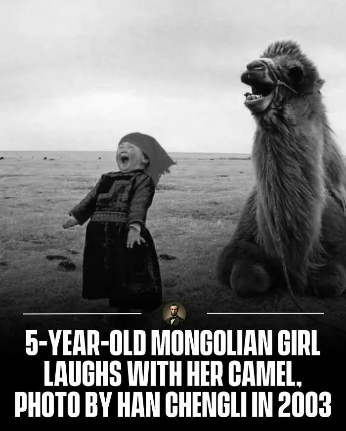 A photo from history of a young Mongolian girl laughing alongside a camel in a grassy field