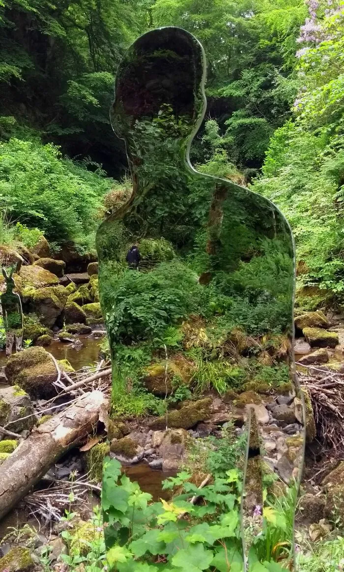 Reflective human sculpture blending into green forest near a stream