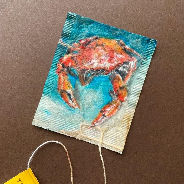 A detailed miniature painting of an orange crab on a used tea bag.