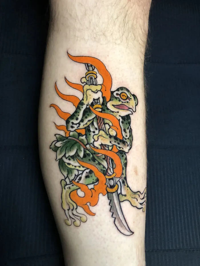 Traditional style tattoo of frog holding a katana with orange flames
