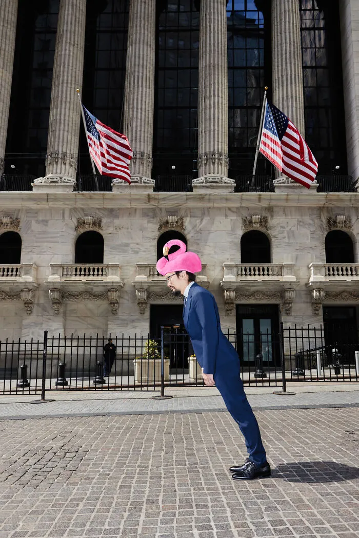 Surreal photo of man in suit wearing pink flamingo hat standing in front of large building with American flags