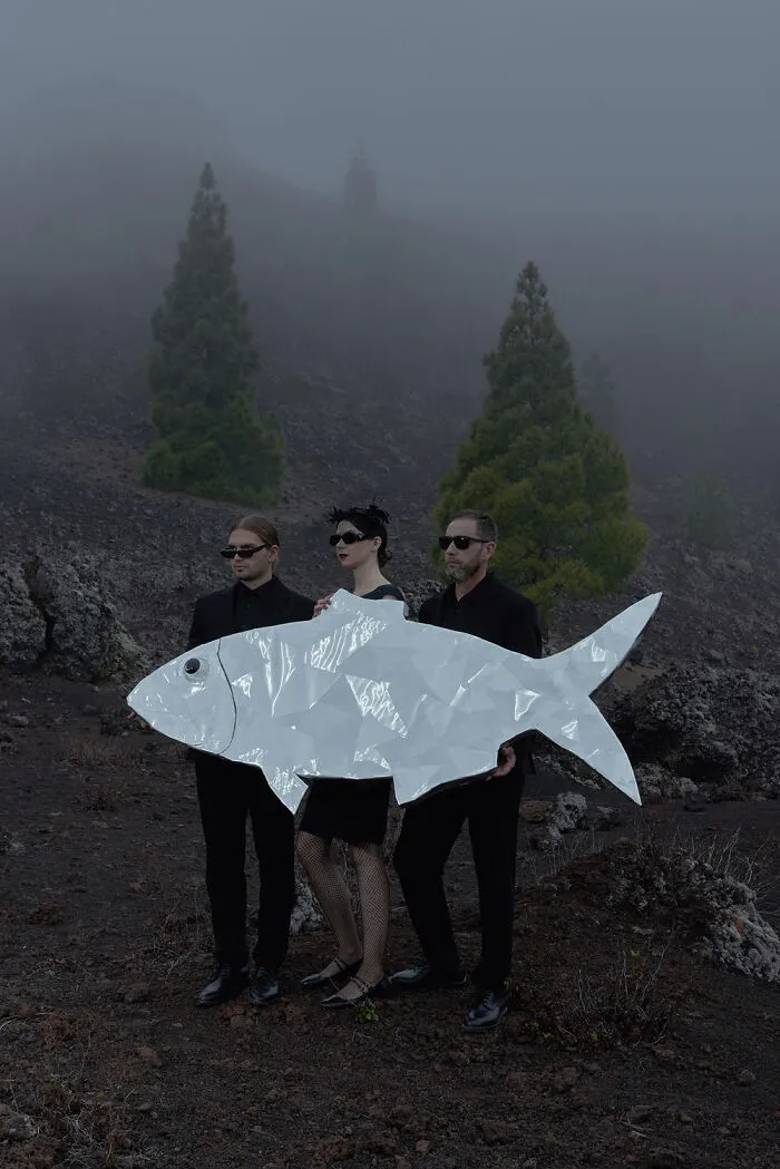 Surreal photo of three people in black holding a large silver fish-shaped object in foggy landscape