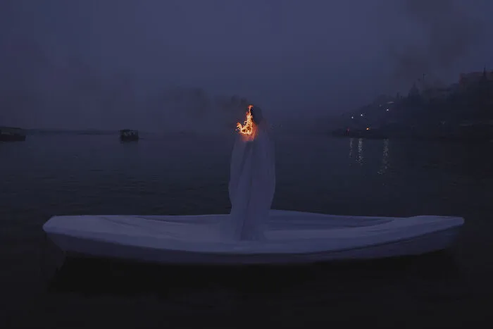 Surreal art expedition photo of figure wrapped in white on boat at night with flame