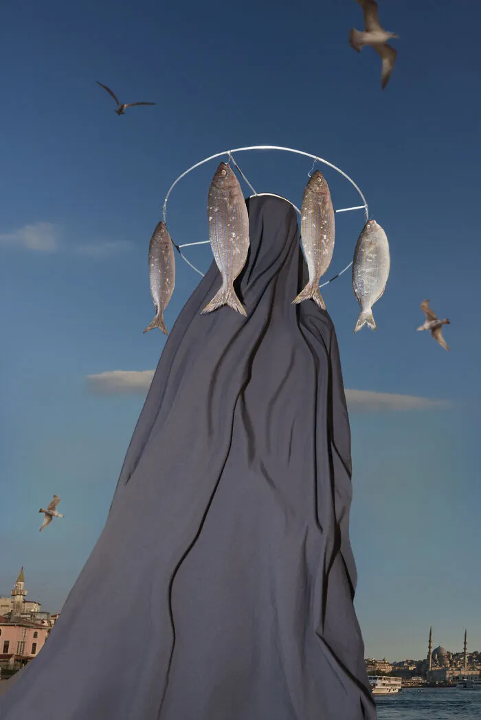Surreal figure draped in fabric with fish hanging from halo against sky