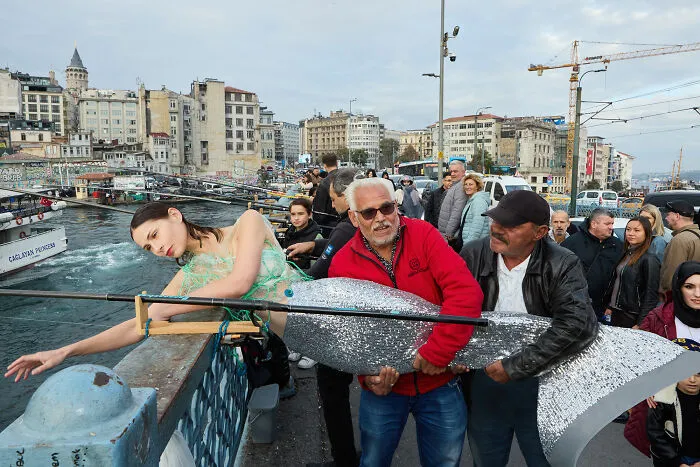 Surreal art expedition photo of model posing with giant sparkling fish sculpture on busy urban pier