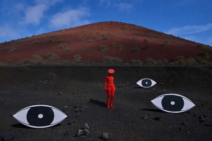 Surreal art expedition image of person in red with large eye cutouts on dark volcanic ground