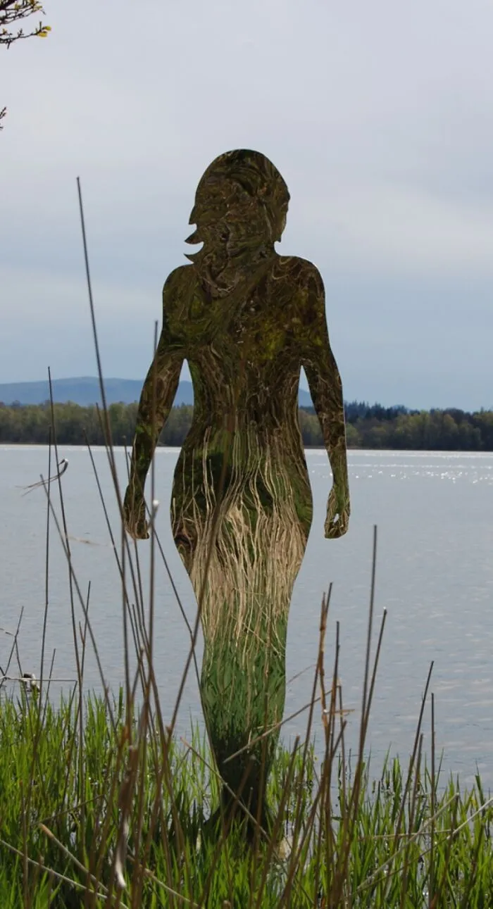 Reflective human silhouette sculpture blending with lakeside reeds and distant trees