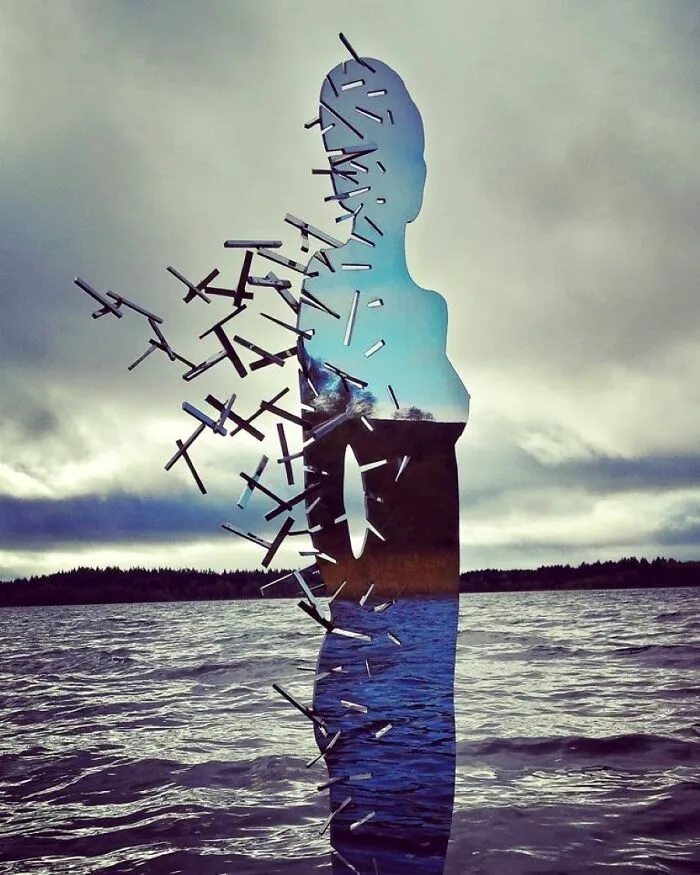 Reflective sculpture of human figure dissolving into fragments by a lake under cloudy sky