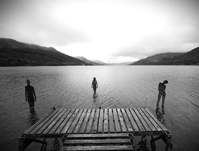 Black and white image of reflective sculptures standing in lake near wooden pier