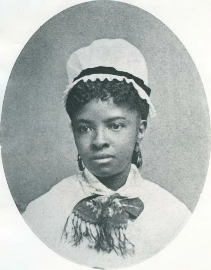 A young Black woman in a white bonnet with dark curly hair and earrings, captured in an oval portrait, highlighting women in history.