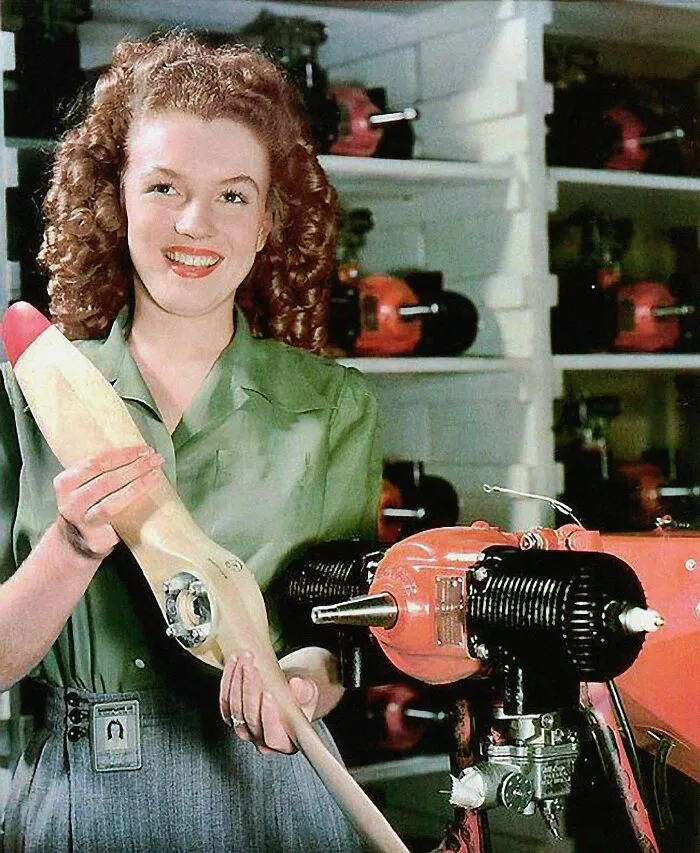 Marilyn Monroe, a prominent woman in history, smiles holding a propeller, with machinery visible in the background.