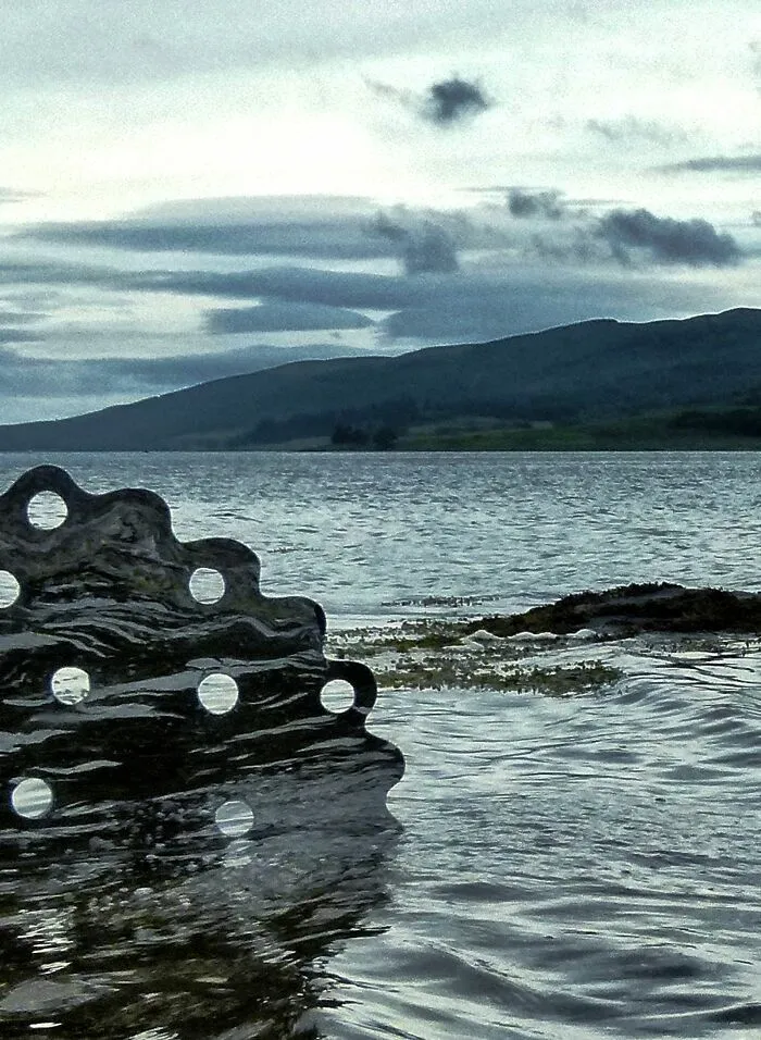 Abstract reflective sculpture with holes partially submerged in water against mountain backdrop