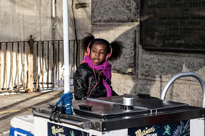 A young girl in pink headphones and a purple scarf singing at a street vendor's cart, an odd coincidence captured by a street photographer.