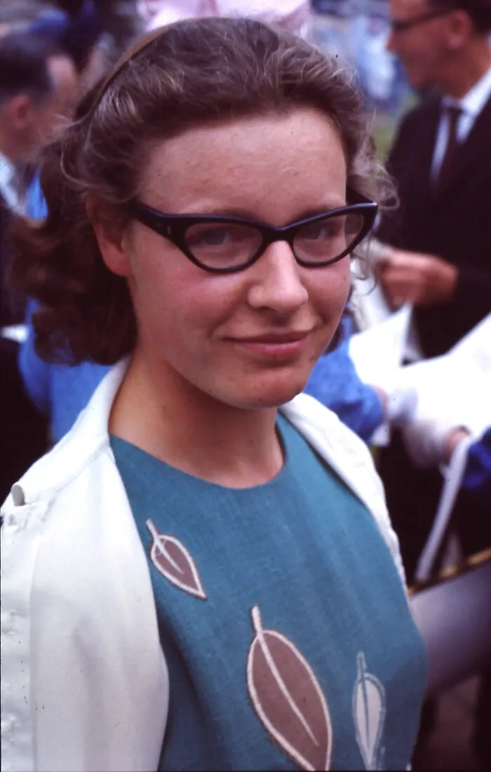 A smiling Woman-In-History wearing glasses, a teal dress, and a white cardigan.