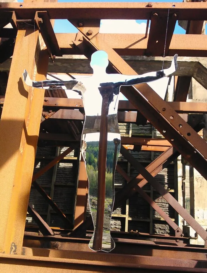 Reflective human sculpture with arms outstretched amid rusted metal framework