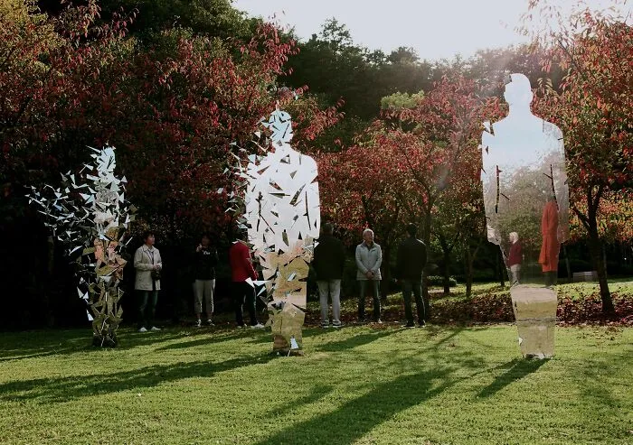 Reflective sculptures of fragmented human figures in park with autumn trees and sunlight