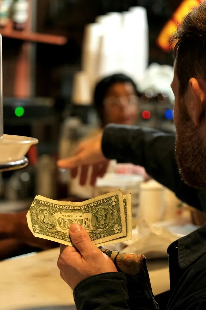 A man holding a one dollar bill at a counter, highlighting cheap tipping habits