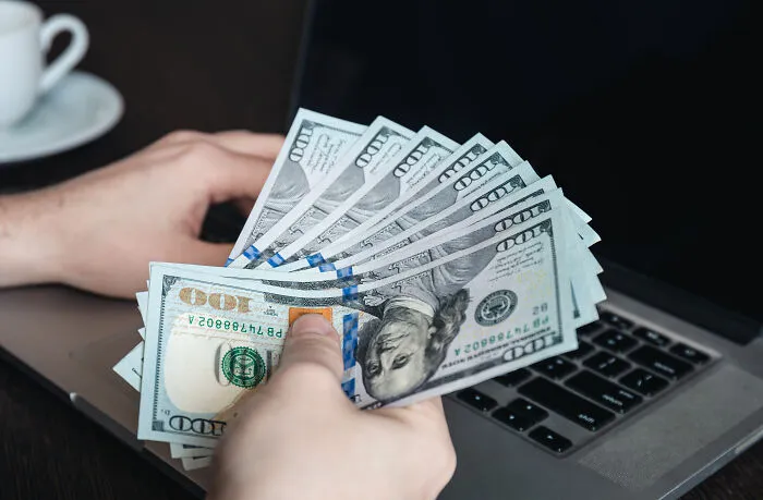 A hand holding US dollar bills in front of a laptop, highlighting marriage regrets