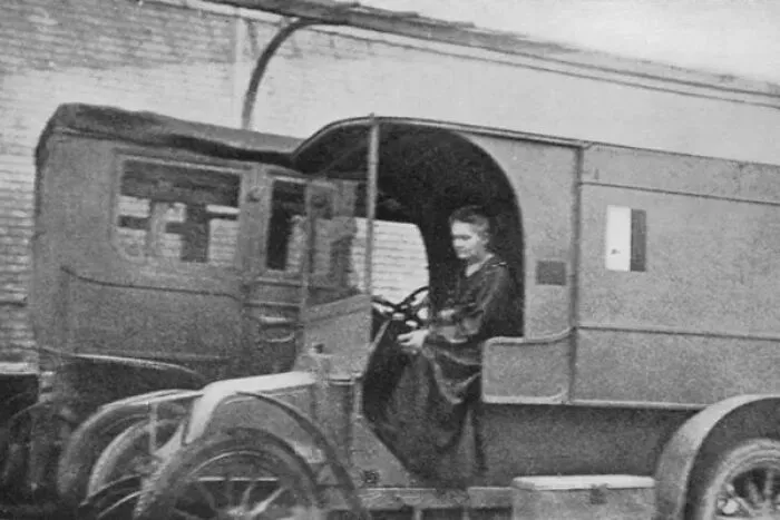 Marie Curie, a notable woman in history, sits in the driver's seat of an early model vehicle, possibly an ambulance.