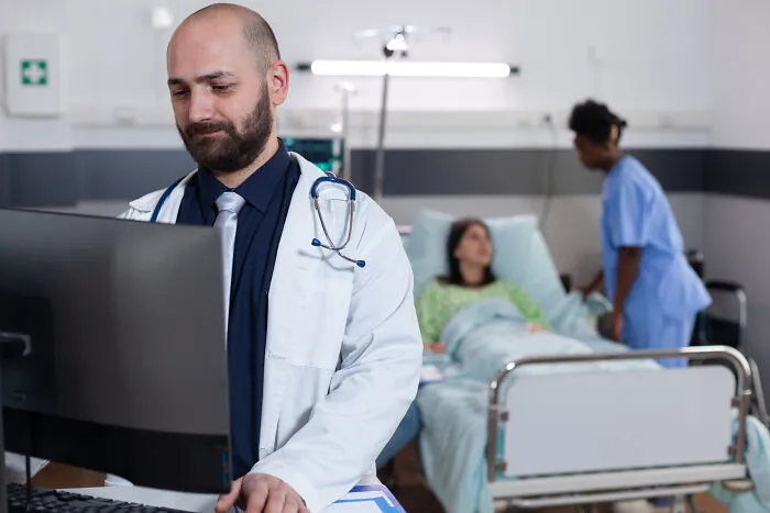 A doctor at a computer, with a nurse and patient in the background, handling pain reactions.