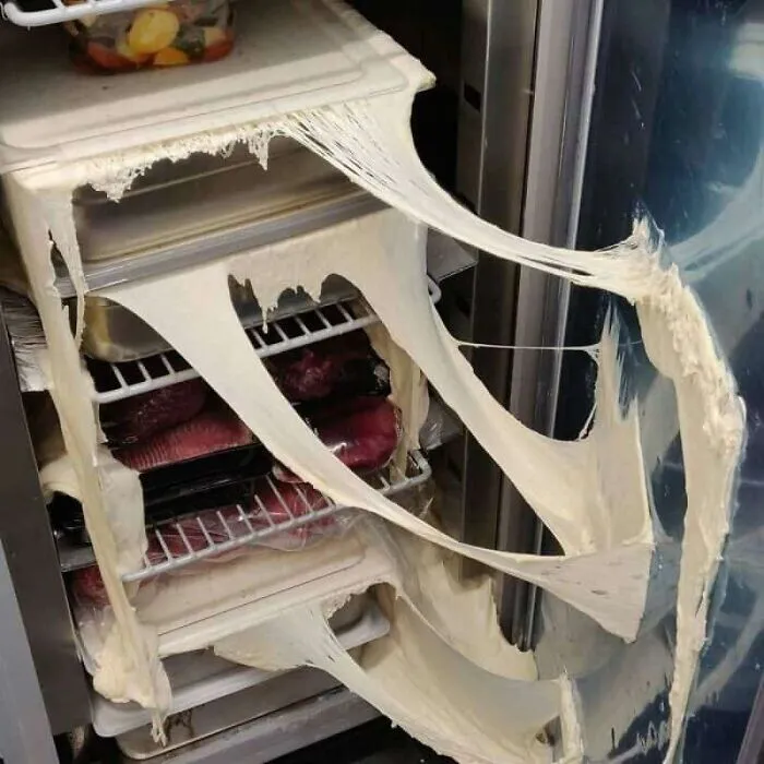 A refrigerator with a severe kitchen fail, showing dough oozing and dripping from multiple shelves.