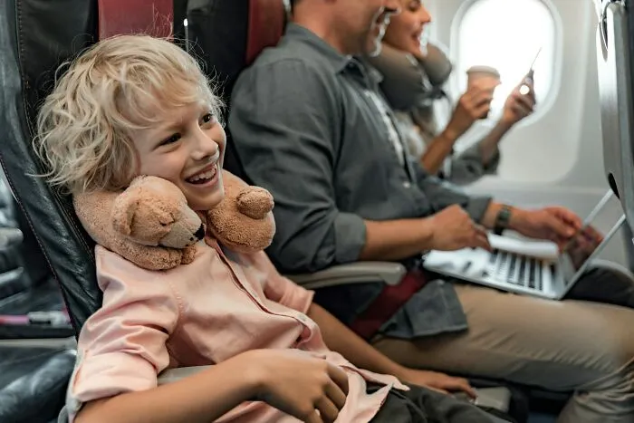A smiling boy with a teddy bear neck pillow sits next to his father on a plane, avoiding a plane seatmate from hell.